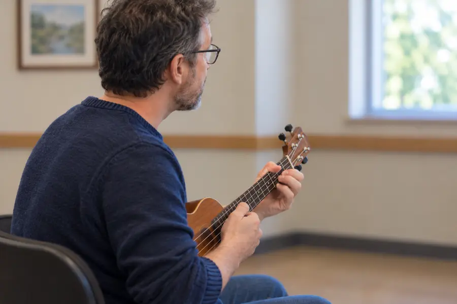 An instructor seated in a bright room, demonstrating a chord on a ukulele.